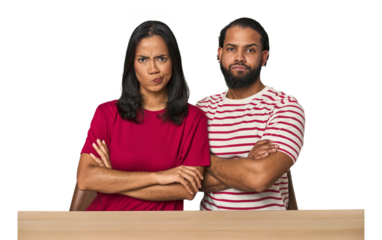 Seated young Latino couple at table frowning face in displeasure, keeps arms folded.