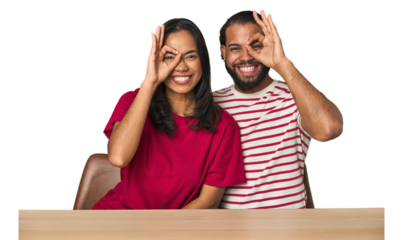 Seated young Latino couple at table excited keeping ok gesture on eye.