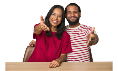 Seated young Latino couple at table stretching hand at camera in greeting gesture.