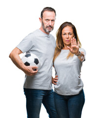 Middle age hispanic couple holding football soccer ball over isolated background with open hand doing stop sign with serious and confident expression, defense gesture