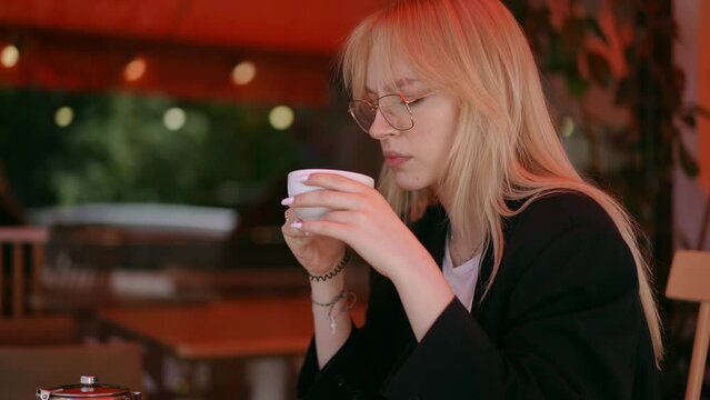 Close-up. A blonde girl with eyeglasses drinks from a porcelain cup, she holds in her hands and cringes with disgust, sitting outdoors at a table on a day on the terrace of a city cafe. The cafe