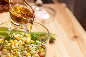 Dressing vegetable salad with vegetable oil. Close-up of hand pouring oil into salad. Olive oil in glass creamer. Vegetable salad in glass saucepan.