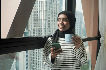 A woman is smiling and holding a cup of coffee while holding a cell phone. Concept of relaxation...