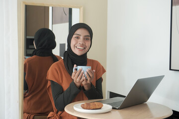 A woman wearing a black scarf and a white striped shirt is sitting at a table with a laptop and a cup of coffee. She is smiling and she is enjoying her coffee and laptop