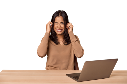 Filipino woman at desk with laptop covering ears with hands.