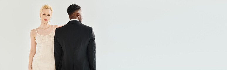 A beautiful blonde bride in a wedding dress and an African American groom standing side by side in a studio against a grey background.