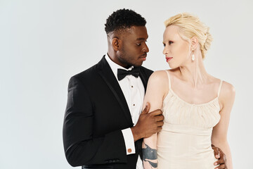 A beautiful blonde bride in a white wedding dress stands next to an African American groom in a classic tuxedo, both in a studio against a grey background.