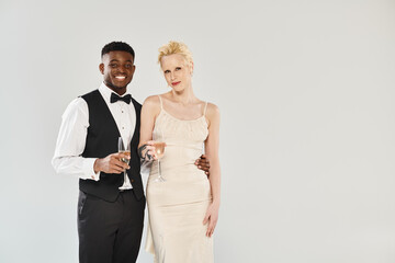 A beautiful blonde bride in a wedding dress and an African American groom in a tuxedo pose elegantly in a studio on a grey background.