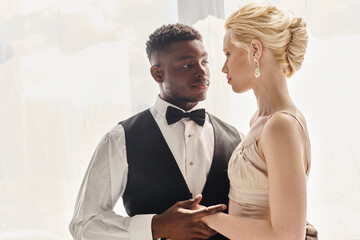 A beautiful blonde bride in a wedding dress and an African American groom standing together in a studio against a grey background.