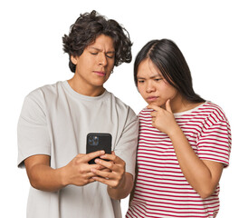 Fototapeta premium Young interracial couple using smartphone looking sideways with doubtful and skeptical expression.