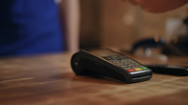 Close-up. An unrecognizable man in a blue apron pushes a bank terminal, that stands on a wooden counter, with his hand and an unrecognizable person holds a bank card to it.