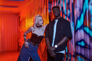 A blonde woman and an African American man stand side by side on an urban street adorned with colorful graffiti walls.