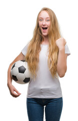 Blonde teenager woman holding soccer football ball screaming proud and celebrating victory and success very excited, cheering emotion