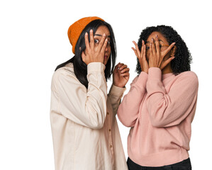 Young Latin friends together in studio blink through fingers frightened and nervous.