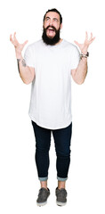 Young hipster man with long hair and beard wearing casual white t-shirt celebrating mad and crazy for success with arms raised and closed eyes screaming excited. Winner concept