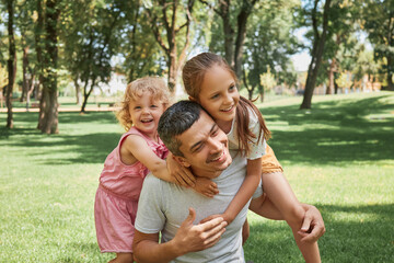 Fototapeta premium Father and daughters love. Kid playing in nature. Happy family outdoors. Cheerful summer park fun. Laughing father carrying his happy joyful daughters on back outdoors