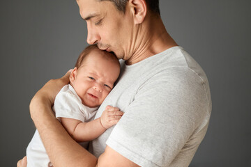 Loving Caucasian father holding his newborn baby in his arms isolated over gray background young man wearing white T-shirt kissing his infant son with love and gentle