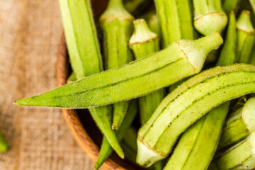 Wooden bowl filled with fresh okra pods placed on a piece of burlap fabric on a rustic wooden white surface.