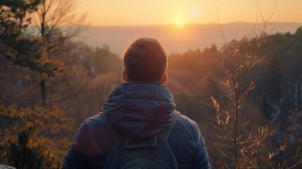 A man is standing on a hill, looking at the sun. He is wearing a blue jacket and a backpack. The sun is setting in the background, creating a beautiful and serene atmosphere