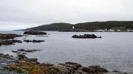 The view from Wester Head spans across the rugged bay of Hant's Harbour, leading to the white lighthouse on the Bay de Verde Peninsula in the Avalon region of Newfoundland, Canada, under a gloomy sky.