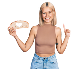 Beautiful young blonde woman holding bread loaf with heart shape smiling with an idea or question pointing finger with happy face, number one