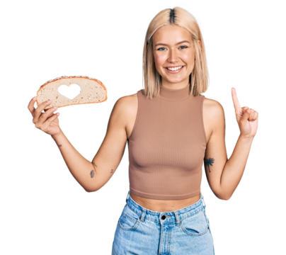 Beautiful young blonde woman holding bread loaf with heart shape smiling with an idea or question pointing finger with happy face, number one - Powered by Adobe
