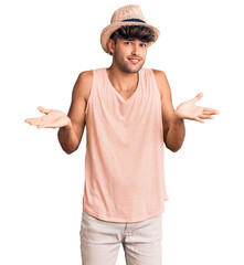 Young hispanic man wearing summer hat clueless and confused expression with arms and hands raised....