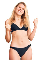 Obraz premium Young beautiful blonde woman wearing bikini very happy and excited doing winner gesture with arms raised, smiling and screaming for success. celebration concept.