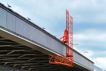 Orange bridge maintenance platform on a granite bridge on a cloudy day. Signifies infrastructure upkeep, engineering, urban maintenance