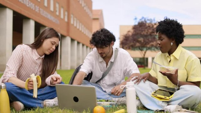 Multiethnic college students working on laptop on campus lawn and eating some food.