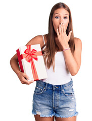 Young beautiful hispanic woman holding gift covering mouth with hand, shocked and afraid for mistake. surprised expression