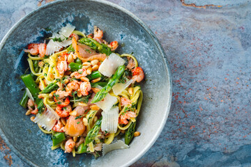 Traditionelle italienische Pasta mit grünem Spargel, Garnelen und geräucherten Lachsstreifen serviert mit Pinienkernen und Parmesan Scheiben als Draufsicht in einer Nordic Design Schale