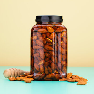 A glass jar filled with almonds and honey is placed on a blue surface, with a few almonds scattered near it and a honey dipper to the left.