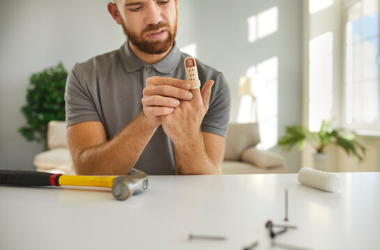 Injured man puts on adjustable finger splint brace after working with hammer at home. Fustrated young bearded man sits at table with hammer and nails. Concept of treatment of injuries at home.