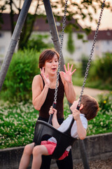 A mother engaging in a playful and animated moment with her young son on a swing at the playground, highlighting joy and togetherness.