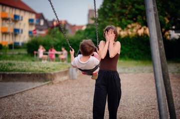 Obraz premium A mother and her young son playing peekaboo while he swings at the playground, sharing a fun and joyful moment together outdoors.