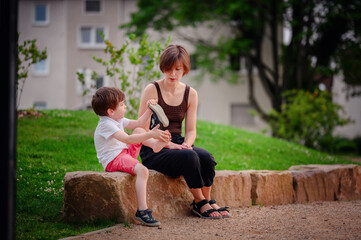 Fototapeta premium A mother and her young son sit together on a large rock in a park. The boy examines his shoe closely while the mother watches, capturing a moment of shared curiosity and care in the outdoors.