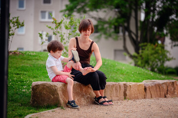 A mother and her young son sit together on a large rock in a park. The boy examines his shoe...