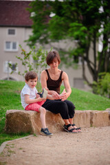Fototapeta premium A mother and her young son sit together on a large rock in a park. The boy examines his shoe closely while the mother watches, capturing a moment of shared curiosity and care in the outdoors.