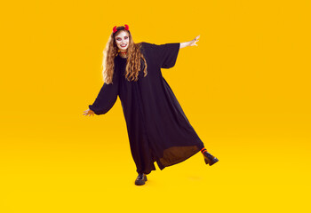 Naklejka premium Woman in Halloween costume moving, dancing, rocking and having fun. Full length shot of happy young girl wearing long black robe and devil horns dancing isolated on yellow color background