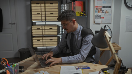 Hispanic detective man working on a laptop in a cluttered police station office.