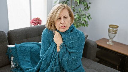 A mature caucasian woman wrapped in a turquoise blanket looks uncomfortable on a gray couch in a...