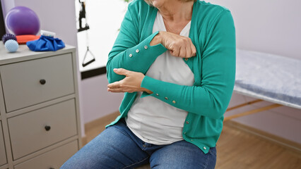 Mature woman holding elbow in pain at a physical therapy clinic, showing signs of discomfort and...