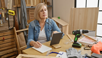 A middle-aged blonde woman ponders over a digital tablet in a cluttered carpentry workshop.