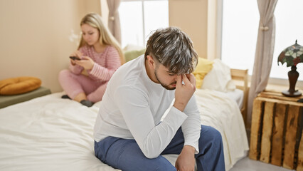 Fototapeta premium A distressed man sits on a bed while a disinterested woman uses a phone, depicting relationship issues in a bedroom setting.