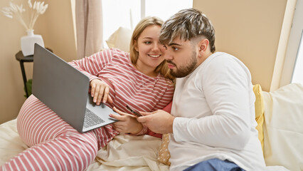 A loving couple, man and woman, shopping online together in a cozy bedroom setting using a laptop and credit card.