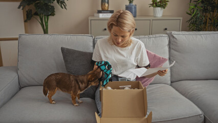 Young blonde caucasian woman sitting in her living room unpacking a box with her chihuahua dog on the couch.