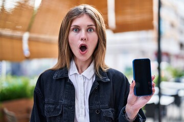 Astonished young blonde woman, mouth agape in disbelief, nervously holding smartphone on bustling...