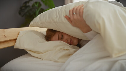 A young caucasian woman covers her head with a pillow while lying in bed in a cozy bedroom setting.