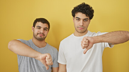 Two hispanic men giving thumbs down gestures isolated on a yellow background
